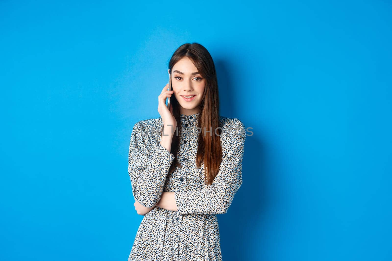 Stylish young woman talking on phone, making takeout order via smartphone, standing in dress against blue background.