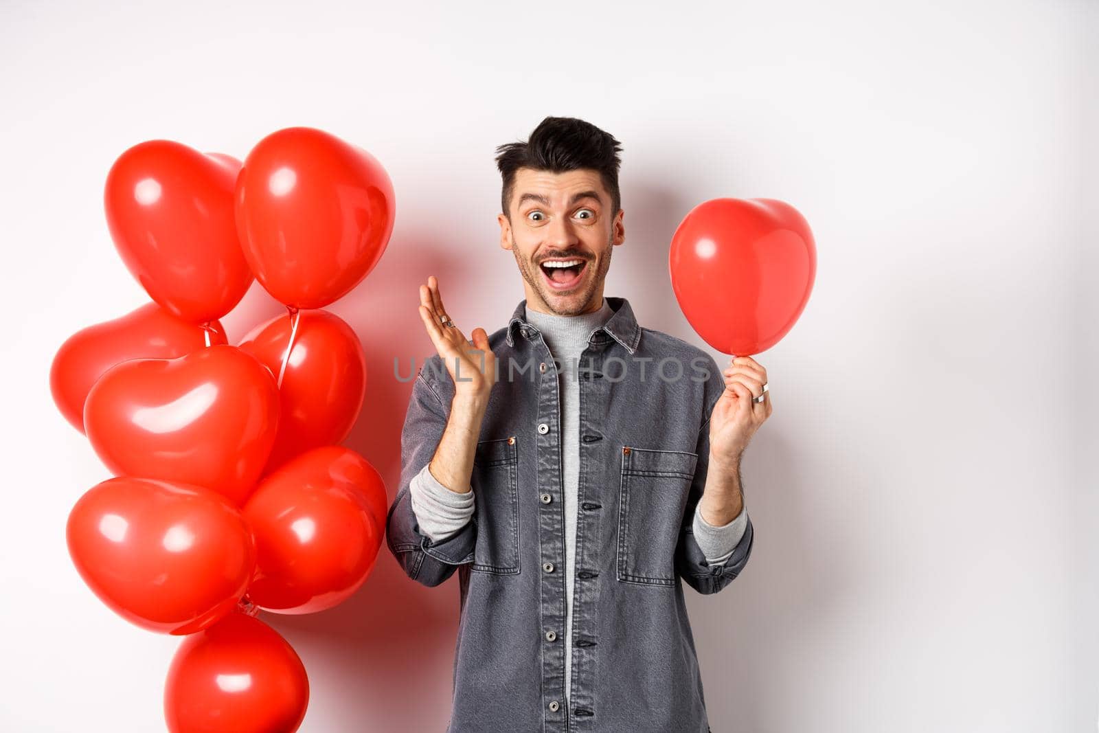 Valentines day concept. Cheerful young man celebrating love holiday, standing near red heart balloons and looking surprised, scream of joy, white background.