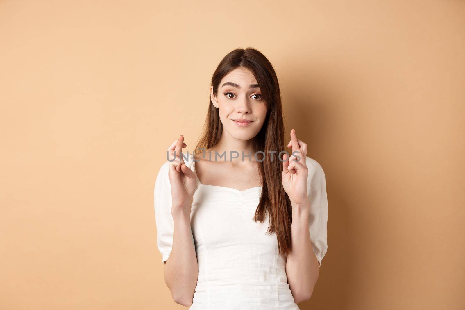 Hopeful tender girl in white blouse making wish, holding fingers crossed and smiling, praying for good luck, waiting for results, beige background.