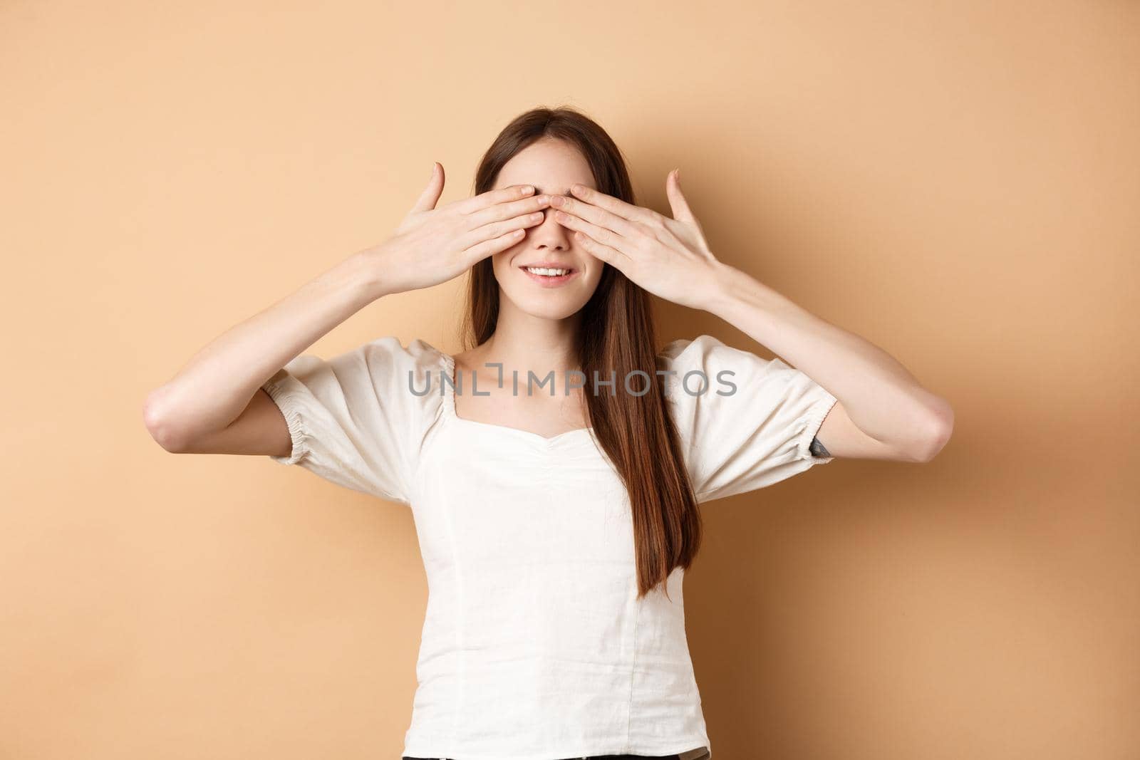 Happy romantic girl covering eyes and waiting for surprise, smiling while anticipating gift, standing on beige background.
