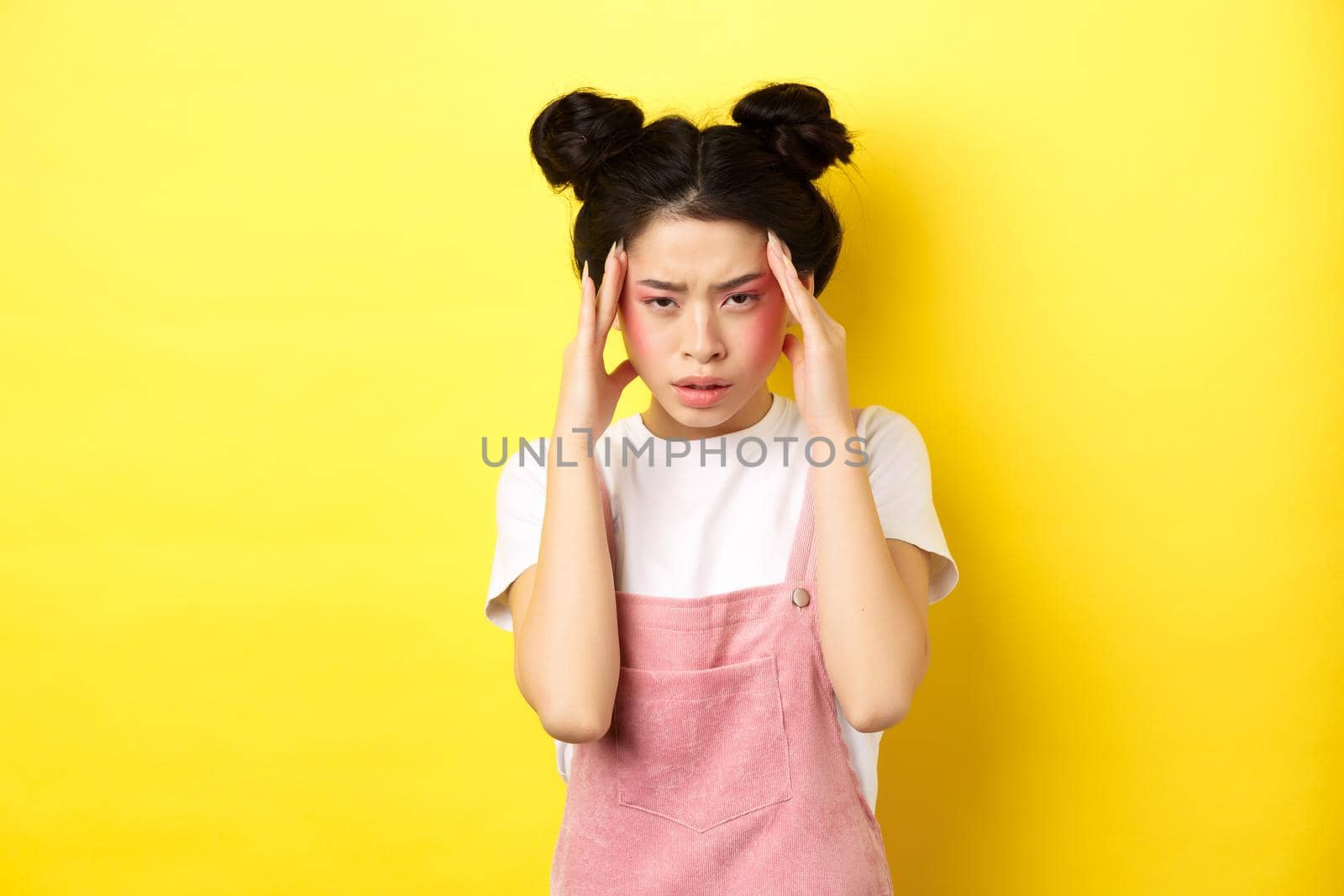 Image of stylish japanese woman with bright makeup, having headache, touching head and looking tired, have painful migraine, yellow background.