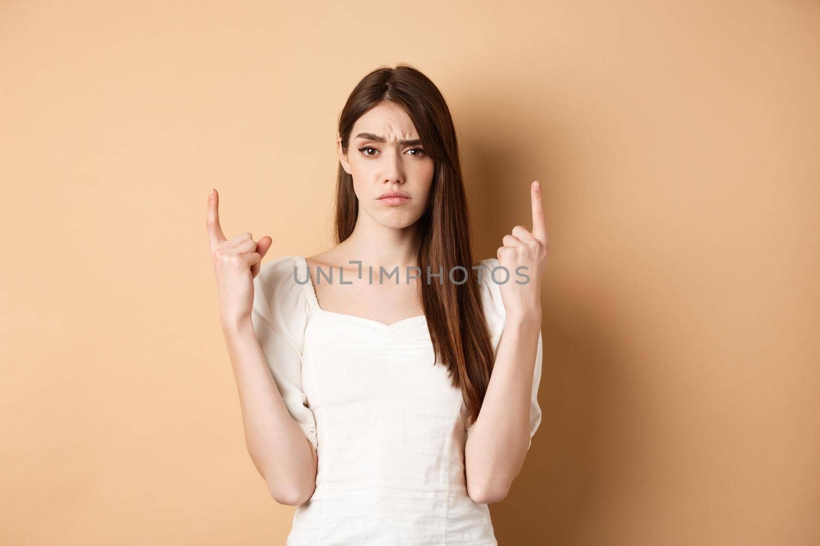 Skeptical woman frowning and pointing fingers up, having doubts about product, standing on beige background. Copy space