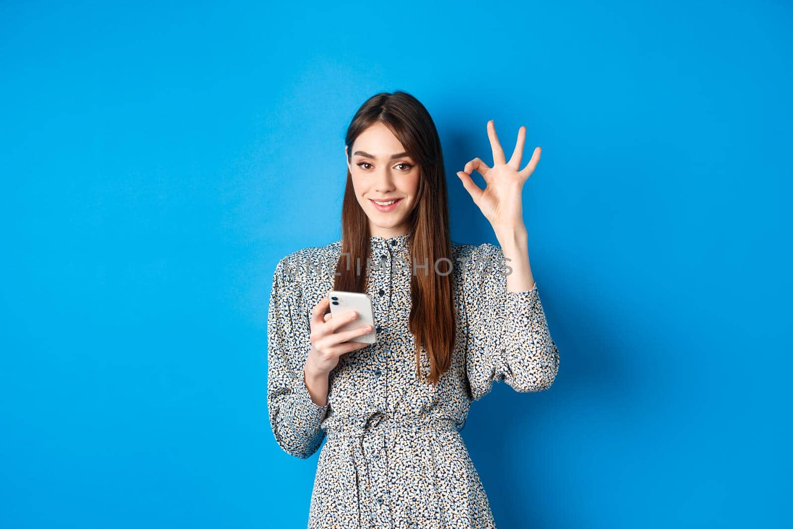 Beautiful young woman in dress showing okay sign and using smartphone, smiling at camera, standing on blue background.