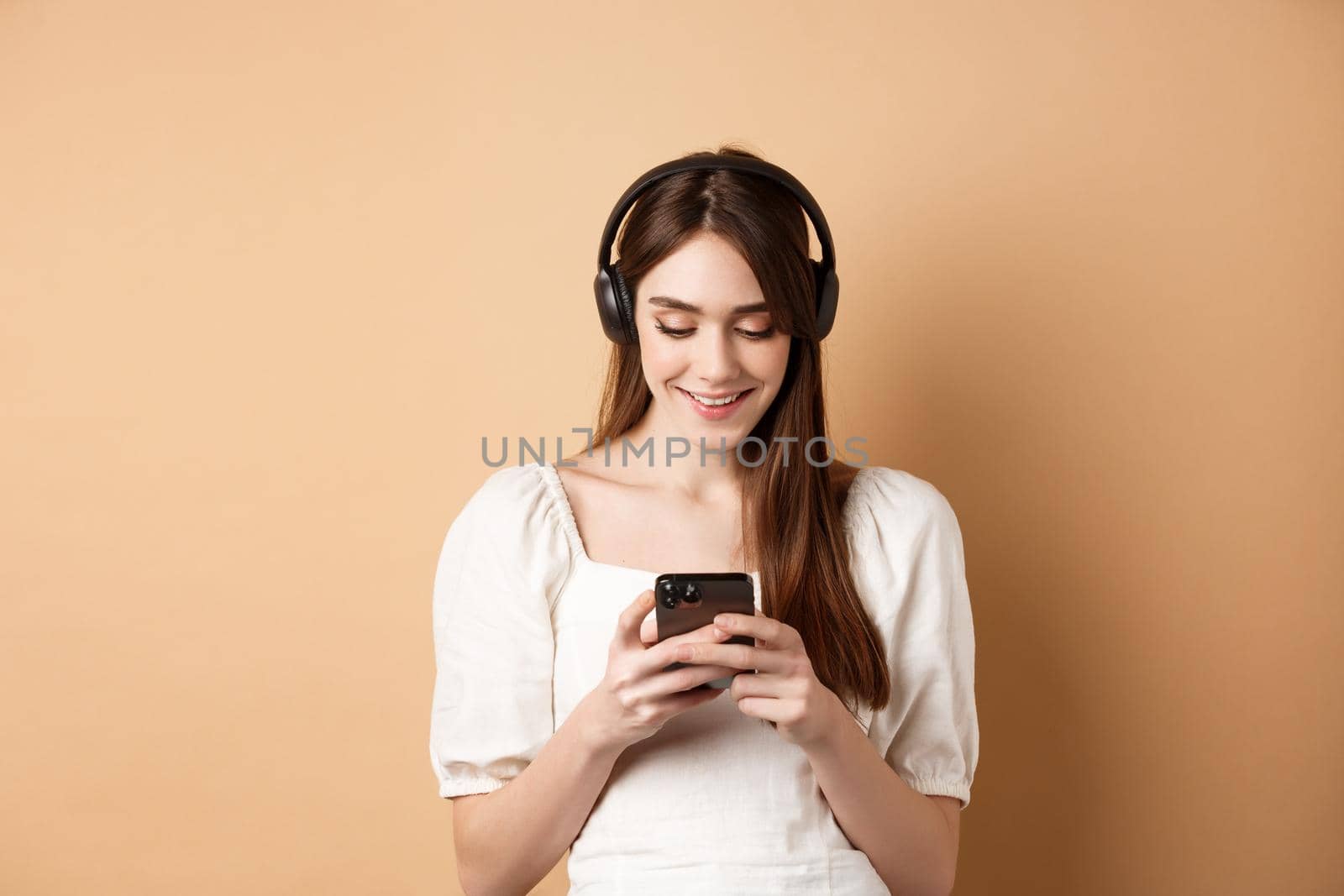 Smiling woman texting message on phone and listening music in wireless headphones, watching video on smartphone, beige background by Benzoix