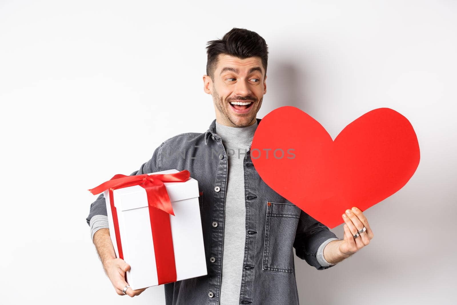 Happy boyfriend making romantic gifts on Valentines day date, holding box with present and big red heart card, smiling cheerful, white background.