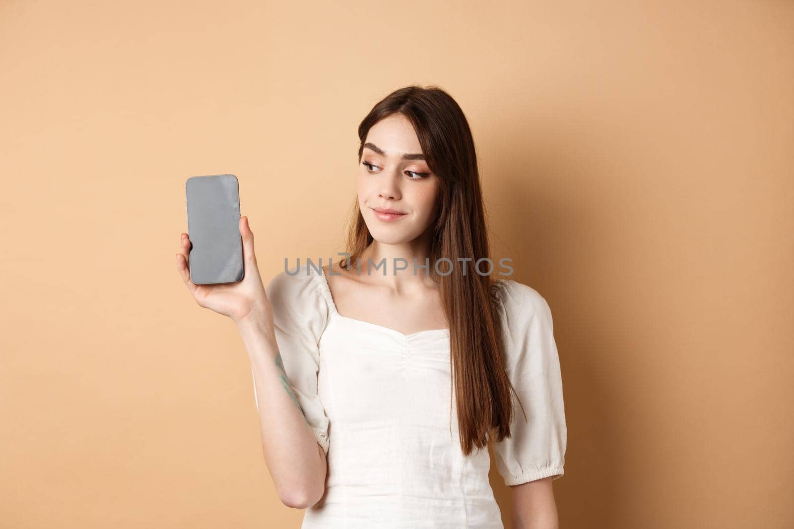 Attractive young woman showing empty smartphone screen, smiling and looking aside, standing on beige background by Benzoix