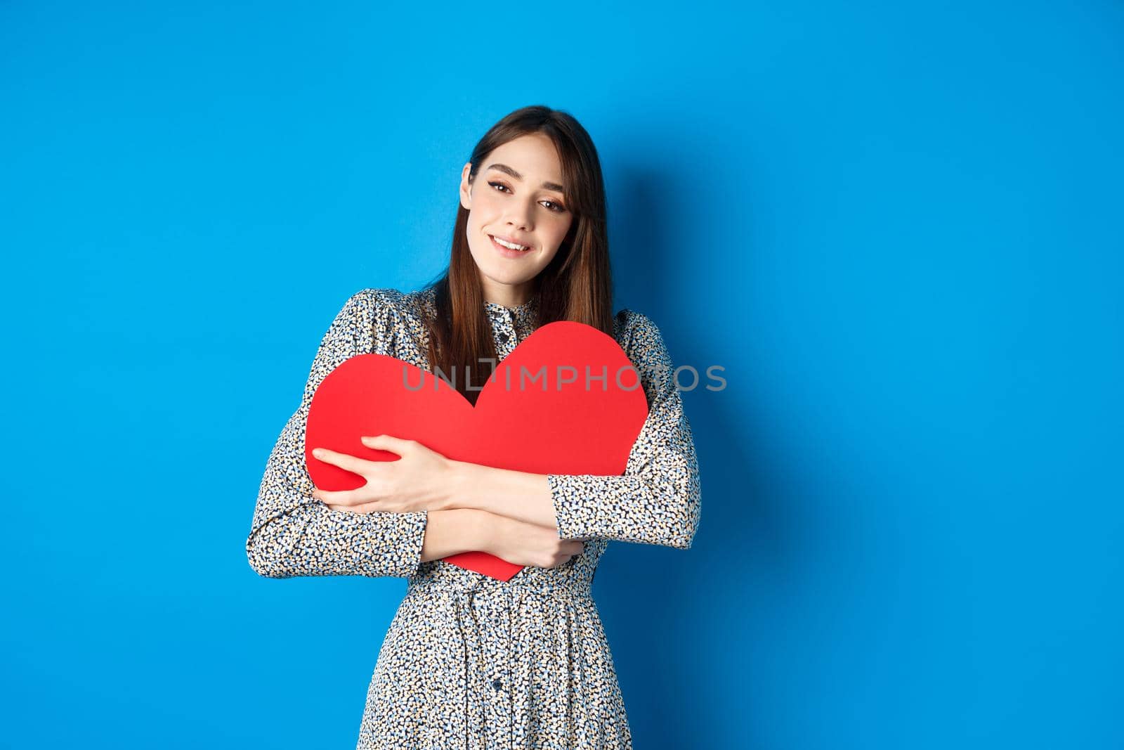 Valentines day. Dreamy romantic woman hugging big red heart cutout, looking sensual at camera, standing in dress on blue background.