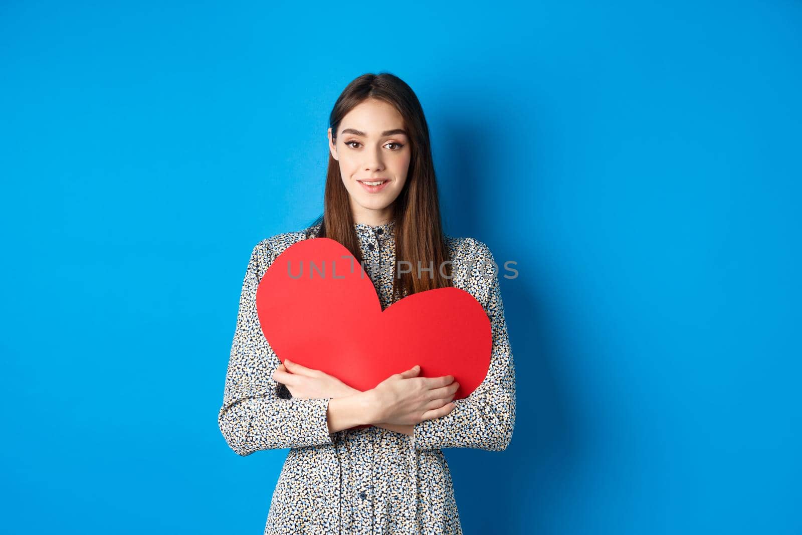 Valentines day. Attractive young woman searching for love, holding big red heart cutout and smiling at camera, standing in dress on blue background.