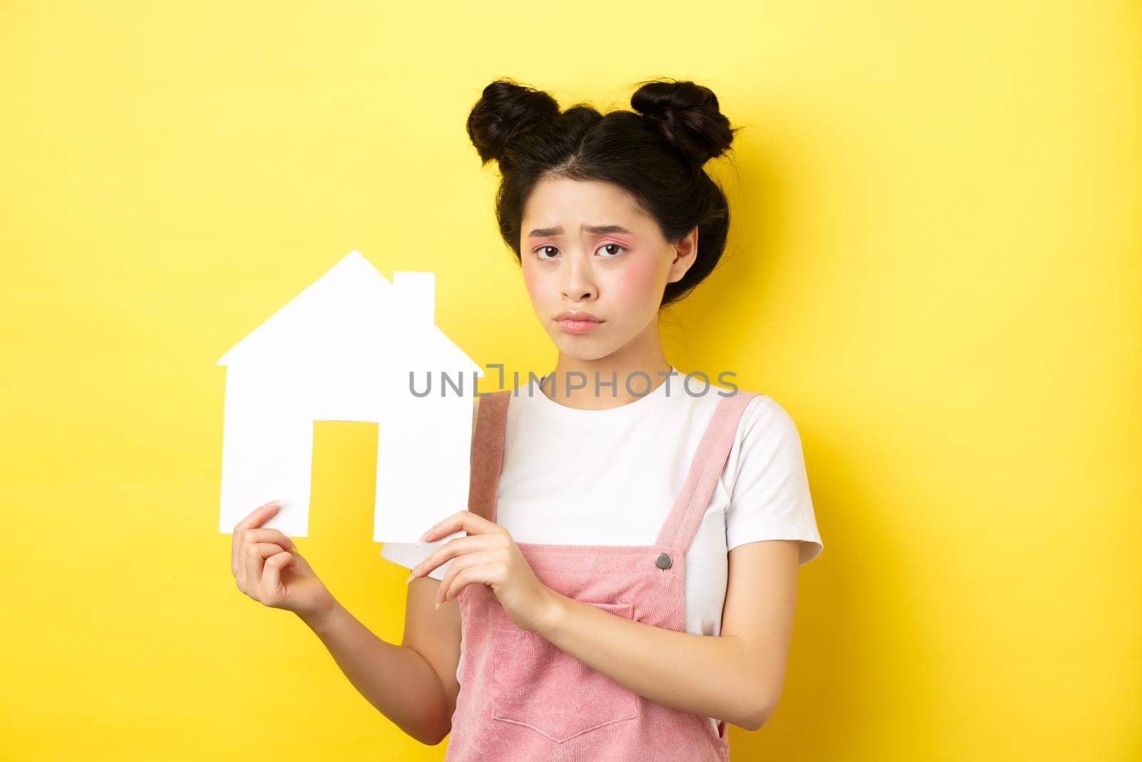 Real estate and family concept. Sad cute asian girl with bright makeup, frowning and feel upset, showing paper house cutout, standing on yellow background.