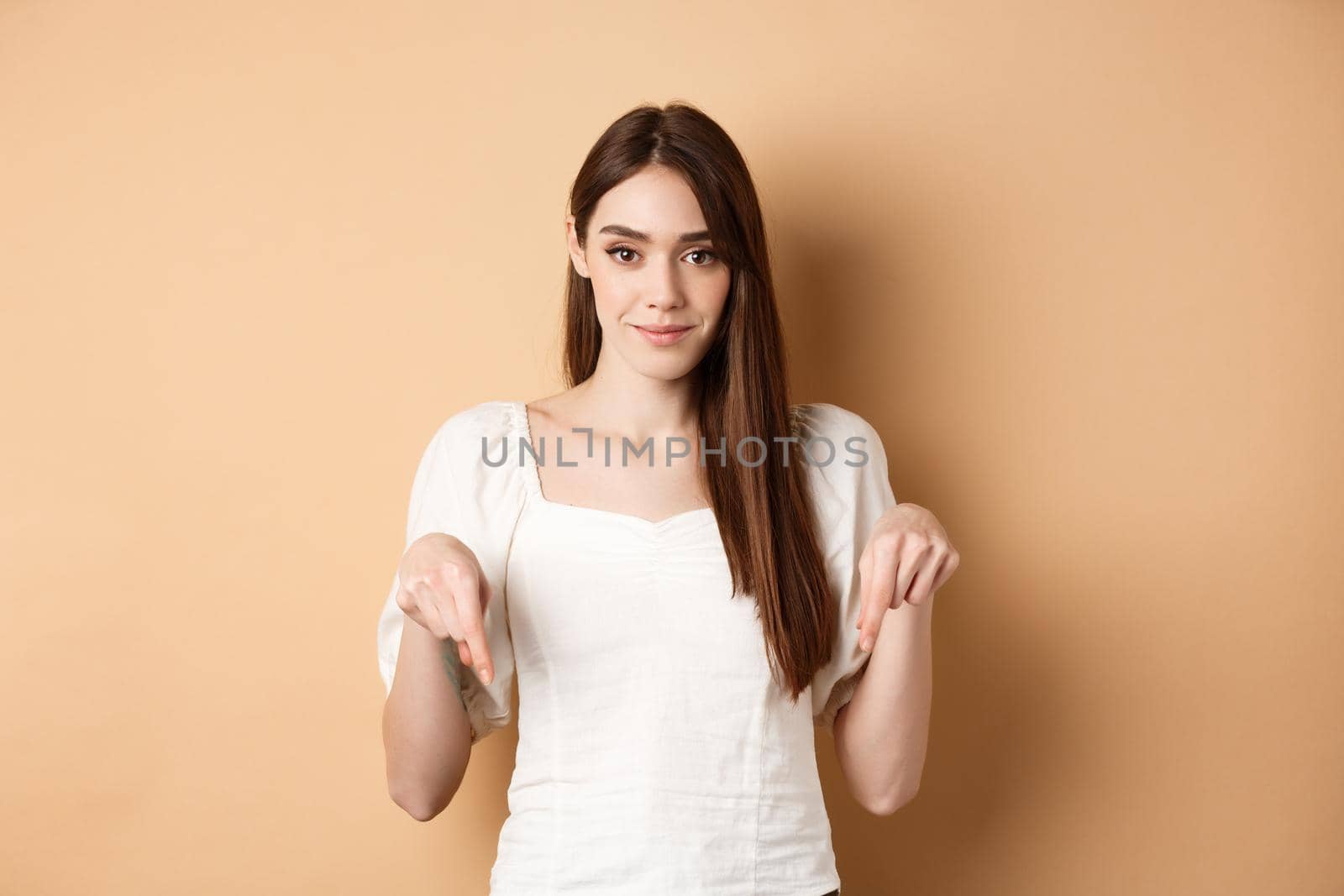 Smiling tender girl in dress pointing fingers down, showing advertisement on empty space, beige background.