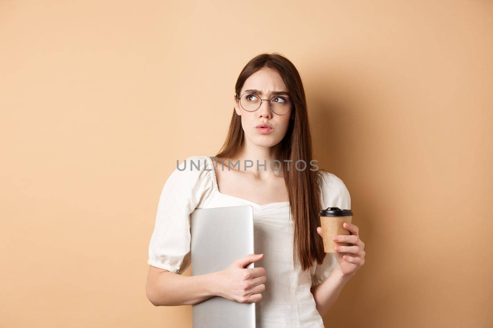 Confused and pensive girl with laptop looking aside suspicious, drinking takeaway coffee, standing against beige background.