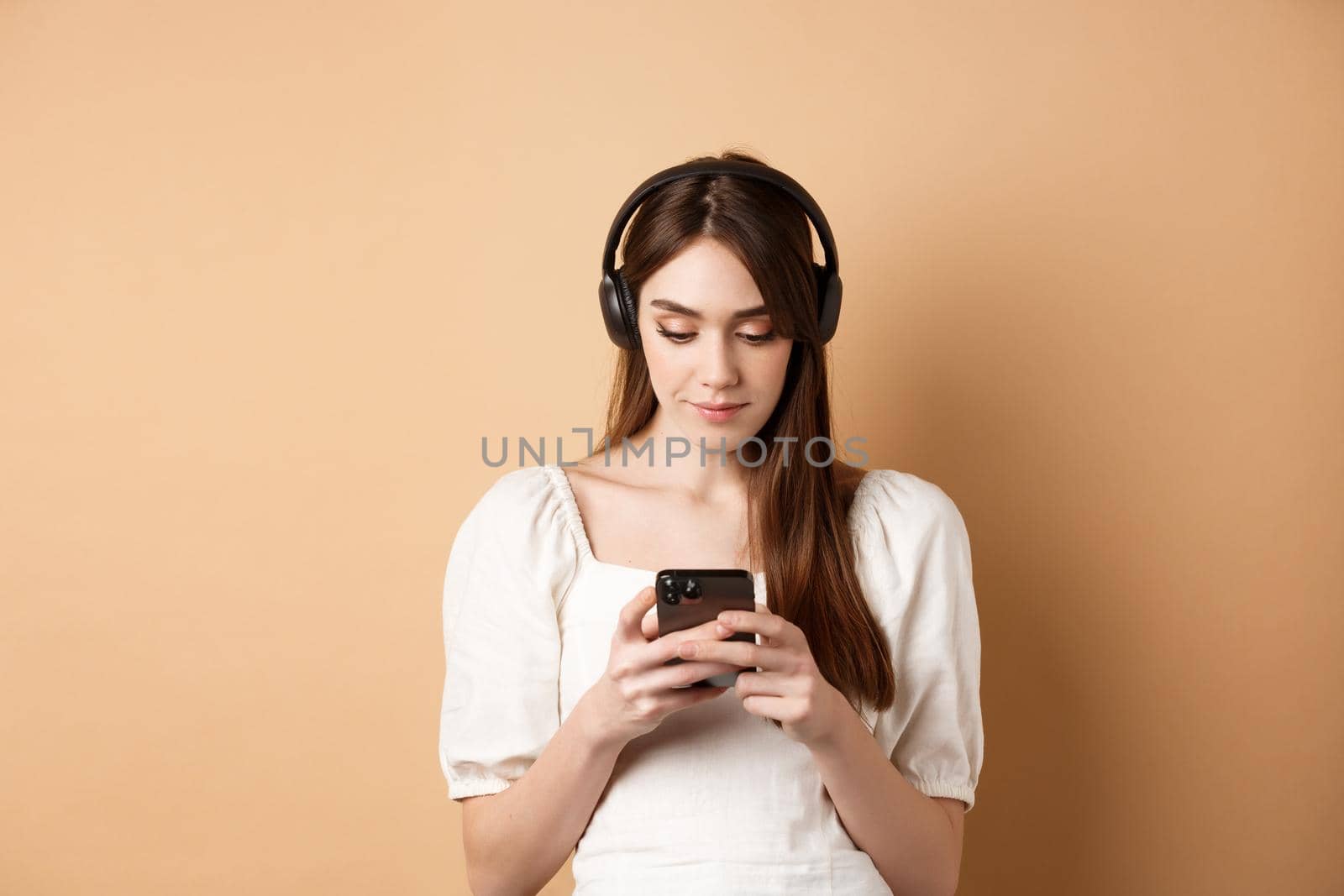 Woman chatting on mobile phone and listening music in wireless earphones, standing on beige background.