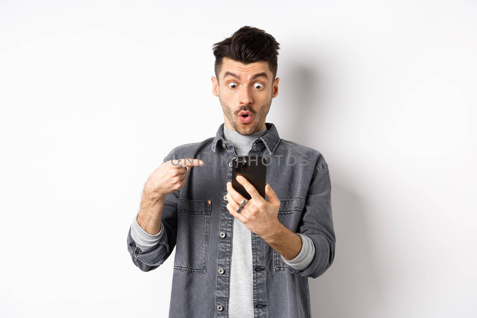 Amazed young man showing cool thing on smartphone, looking and pointing at phone impressed, standing against white background.