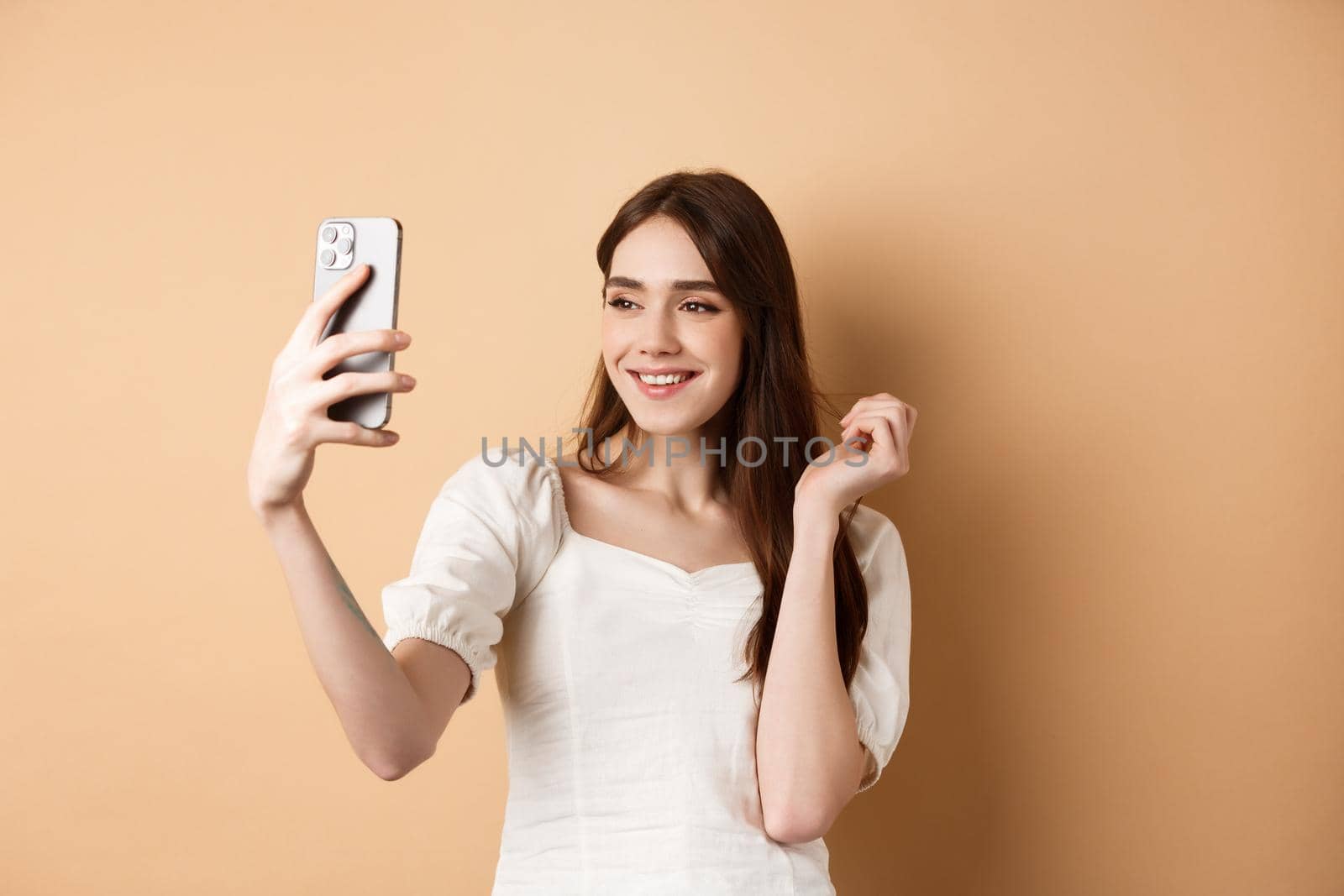 Beautiful woman taking selfie on smartphone, posing for phone photo, standing on beige background.