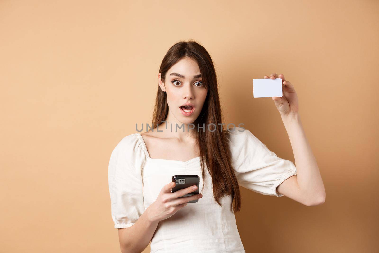 Excited woman showing plastic credit card and using mobile phone app, drop jaw and gasping amazed, checking out bank offer, beige background.