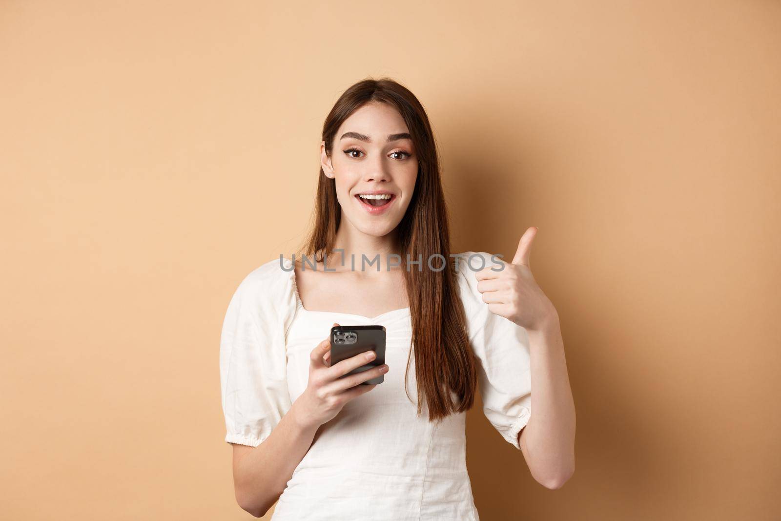 Online shopping concept. Happy young woman showing thumb up and using cellphone, smiling pleased, standing on beige background by Benzoix