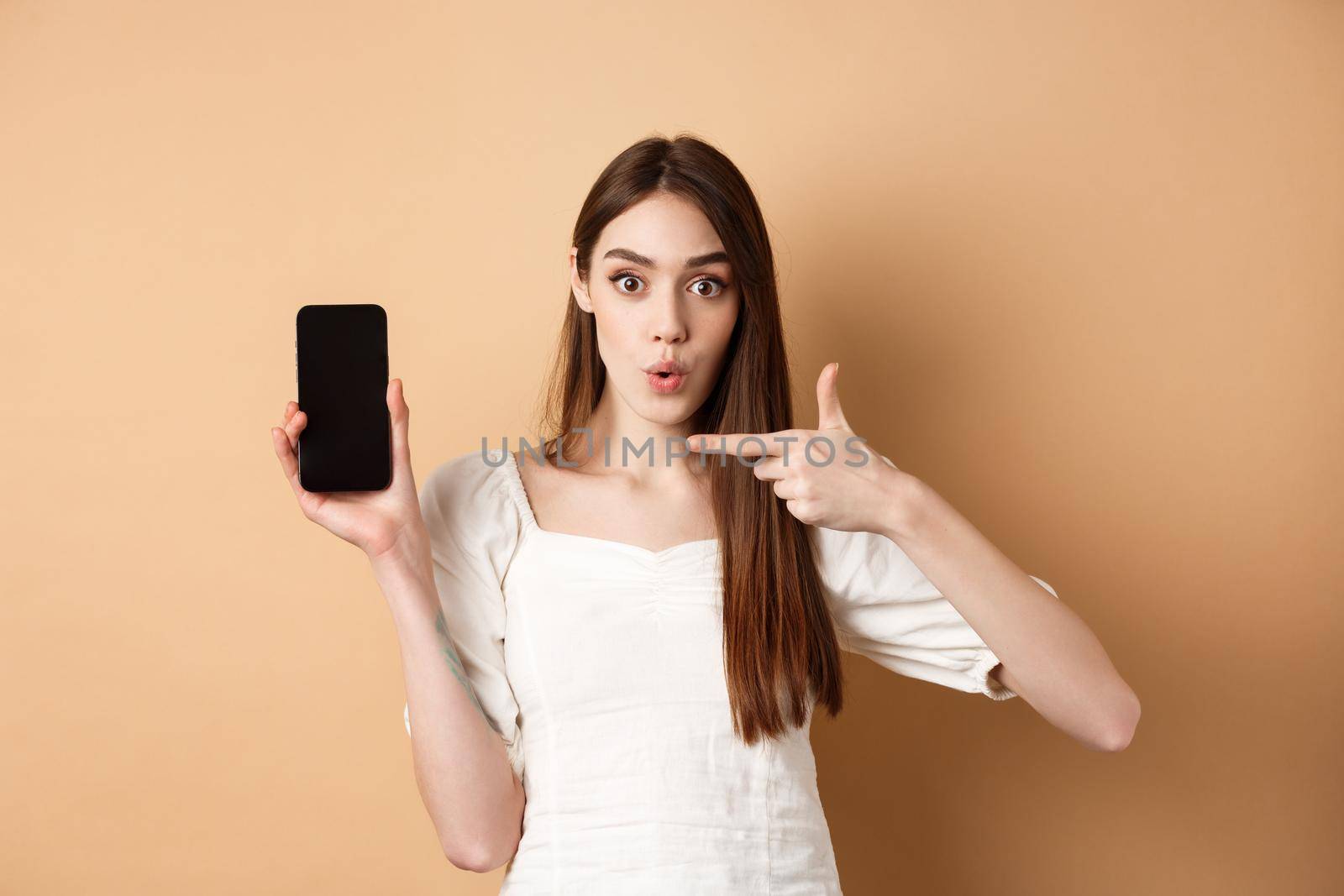 Excited woman showing news on screen, pointing at empty phone and looking surprised, standing on beige background.