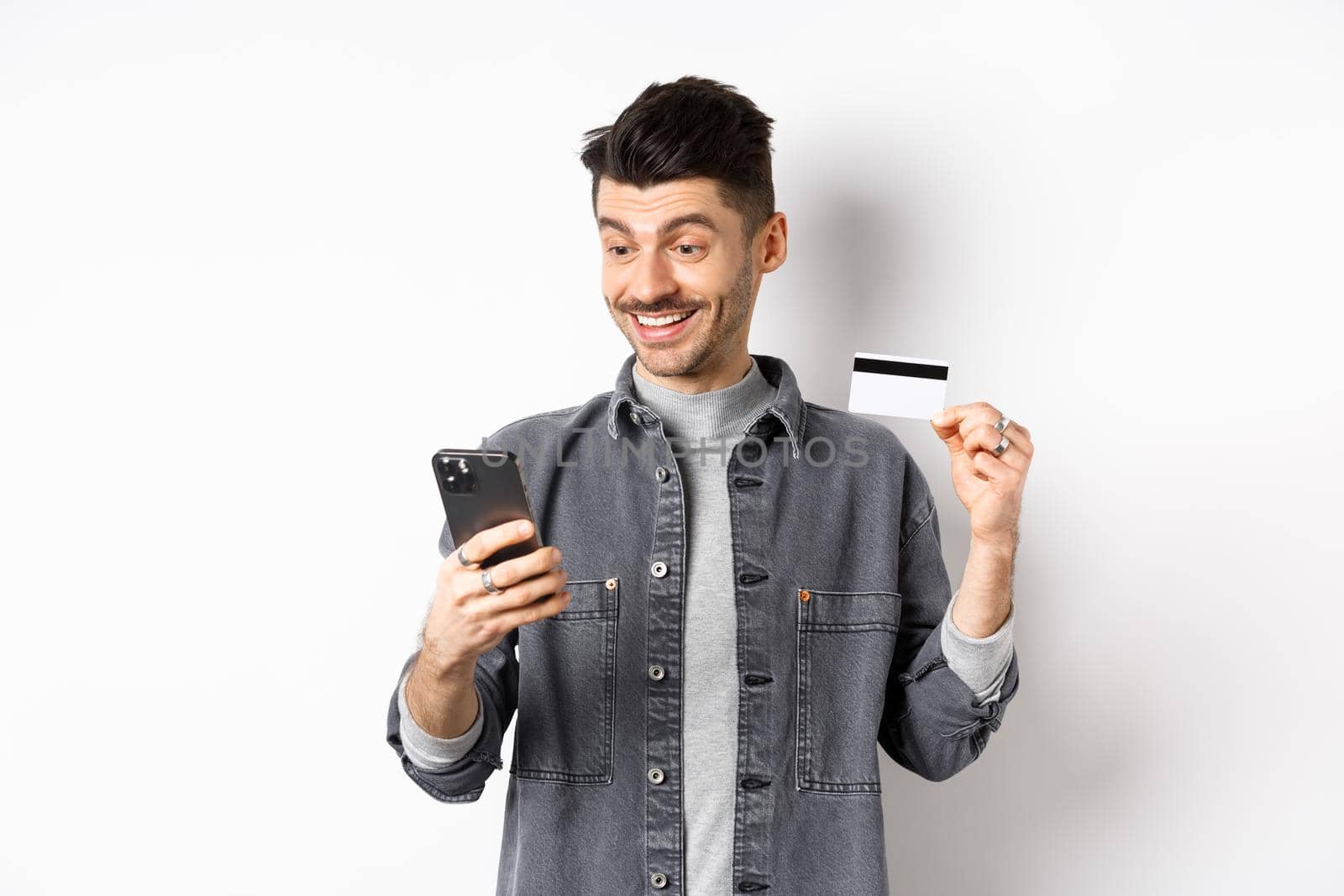 Online shopping concept. Smiling guy paying on mobile phone, showing plastic credit card, buying in internet, white background.