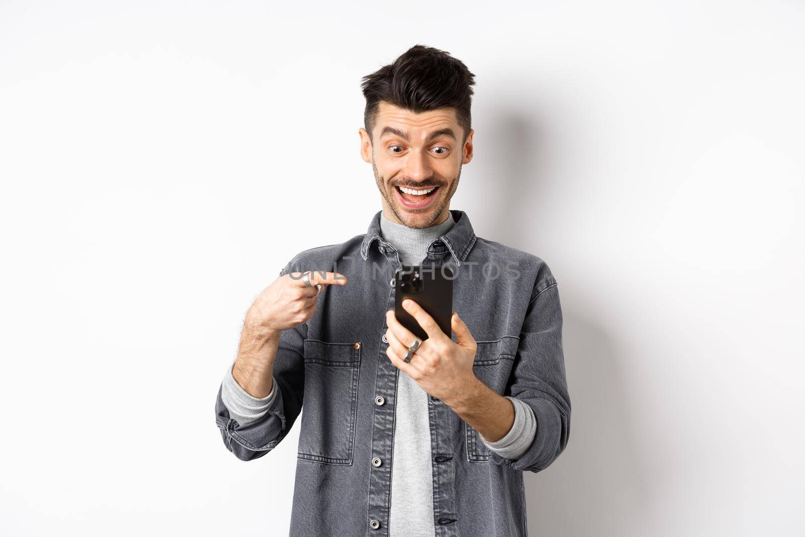 Excited handsome man showing cool news on phone, pointing at smartphone and smiling happy, standing against white background.