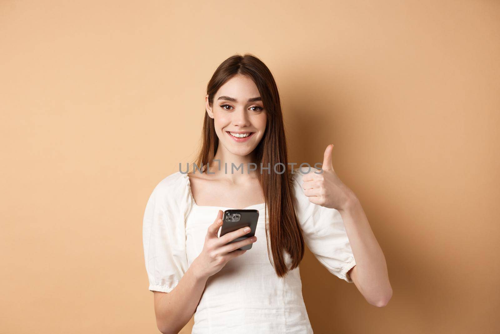 Online shopping concept. Smiling happy woman showing thumb up after using smartphone, holding phone and say yes, standing on beige background by Benzoix