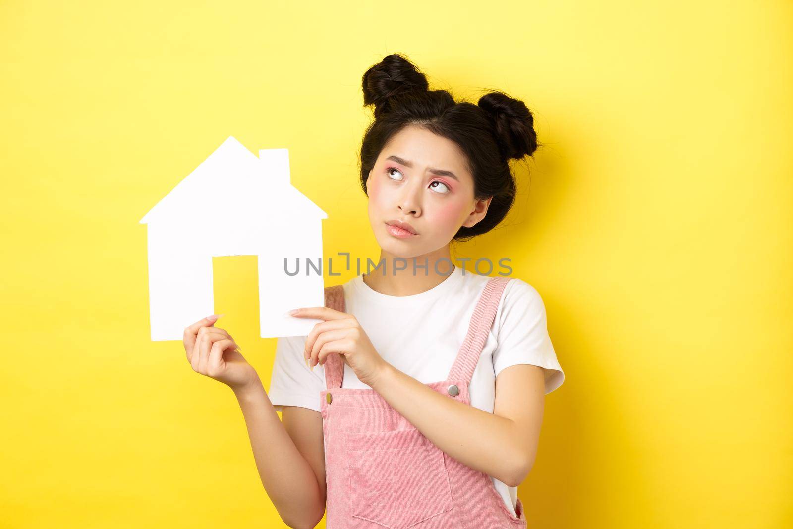 Real estate and family concept. Thoughtful asian girl dream of house, showing paper home cutout and look at upper left corner pensive, yellow background.