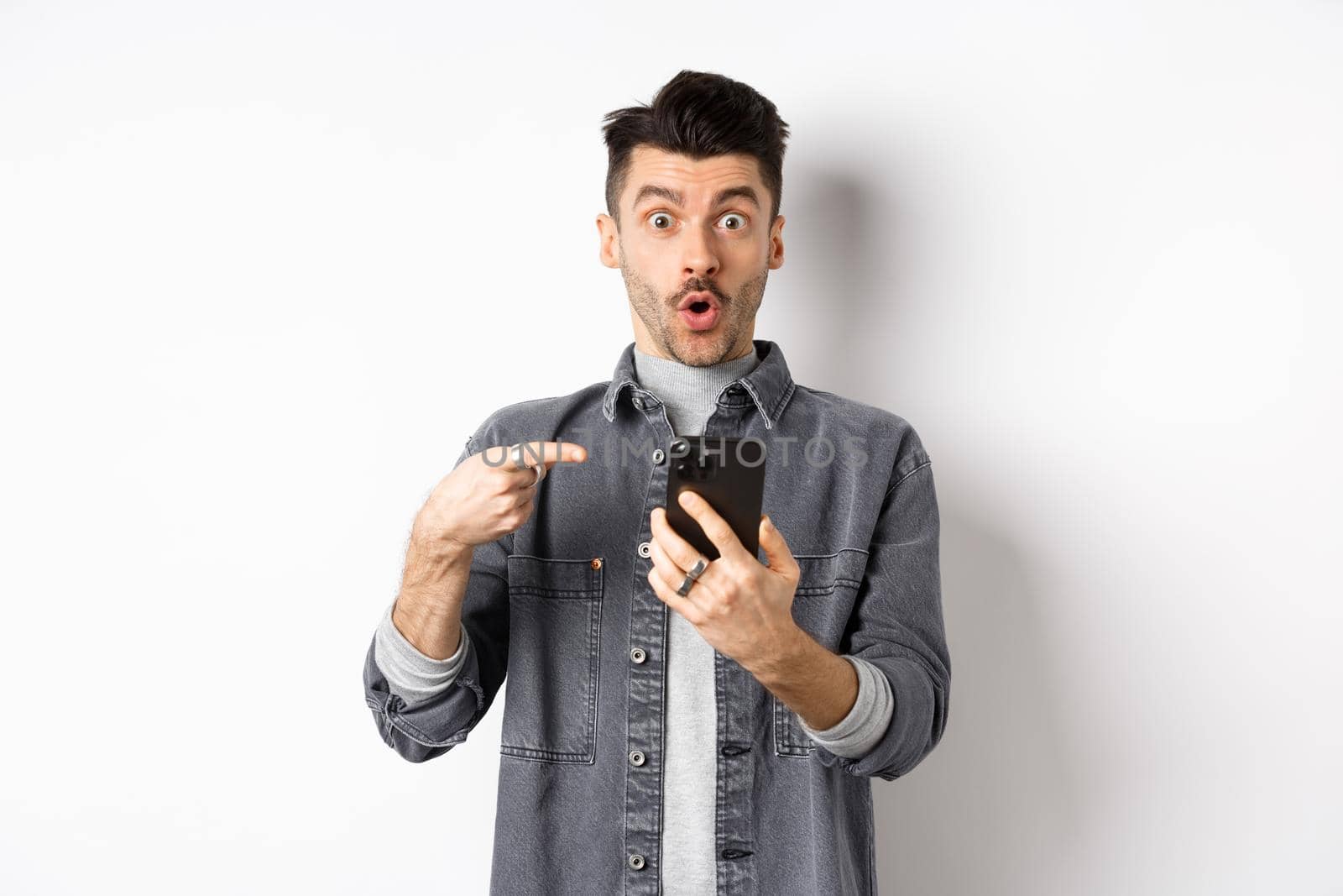 Wow check it out. Excited handsome man showing online offer on phone, pointing at smartphone and looking amazed, standing on white background.