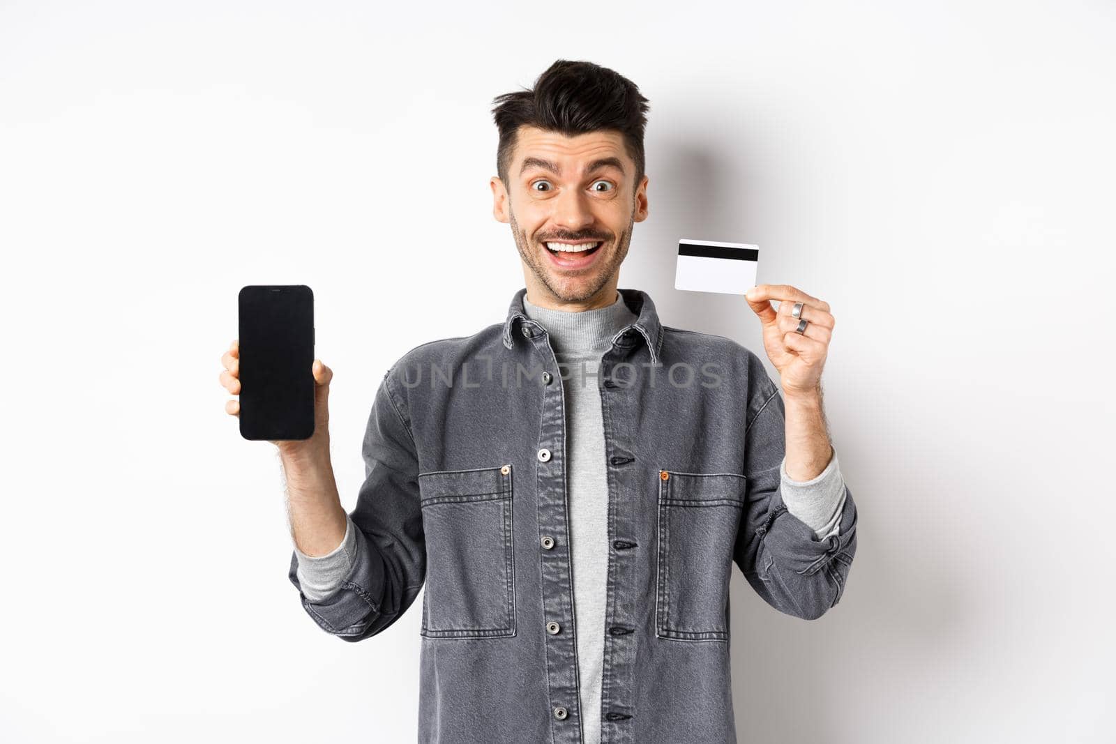 Excited handsome man showing plastic credit card and mobile phone screen, standing on white background.