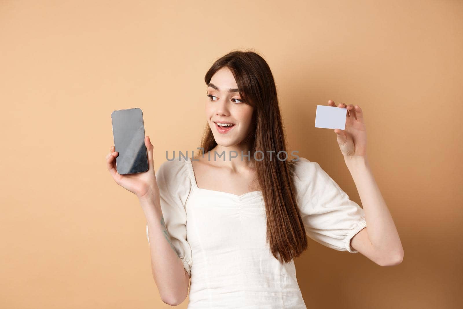 Check this out. Smiling happy young woman showing plastic credit card and empty mobile phone screen, demonstrate application, standing on beige background by Benzoix