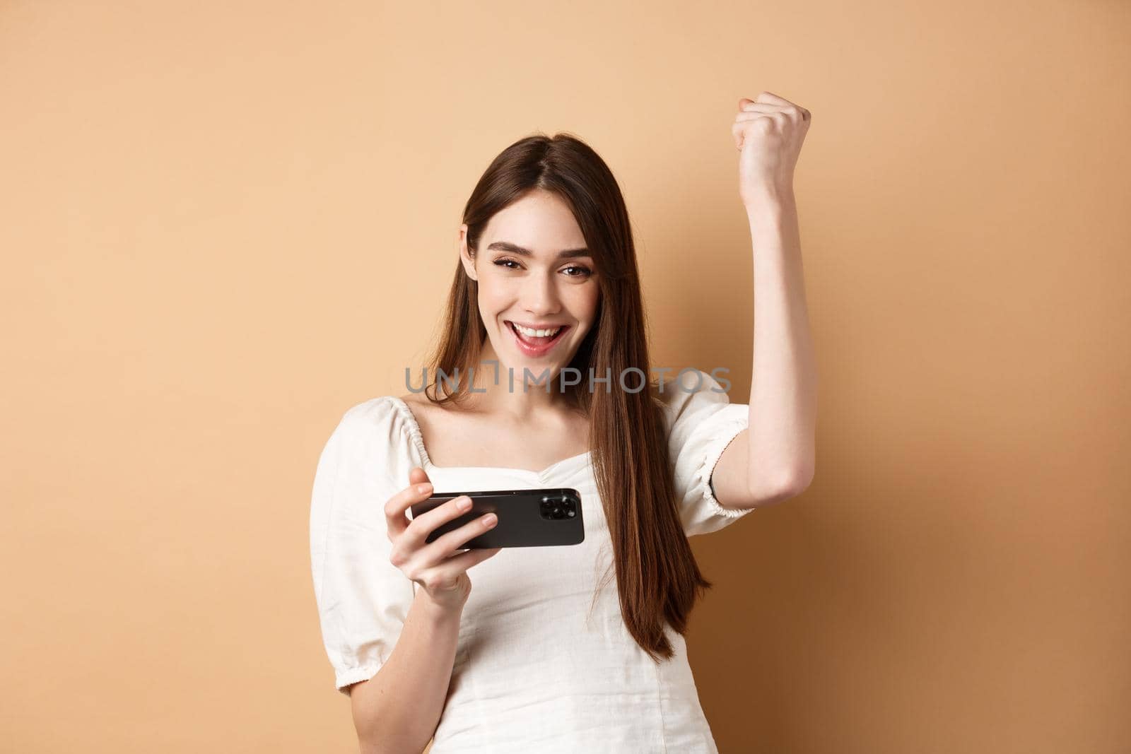Girl winning on mobile phone. Happy woman raising hand up and scream yes with joy, achieve goal in smartphone app, standing on beige background.