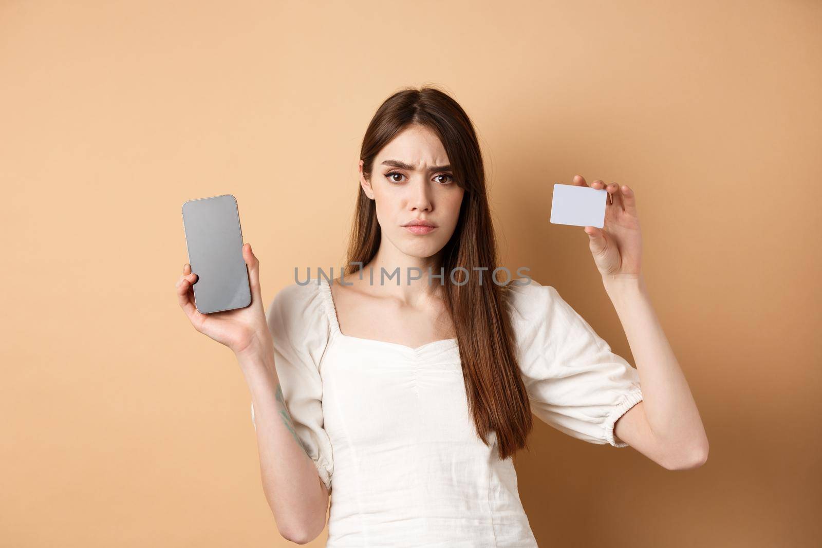 Sad and disappointed girl showing empty smartphone screen and plastic credit card, complaining and frowning, standing on beige background by Benzoix