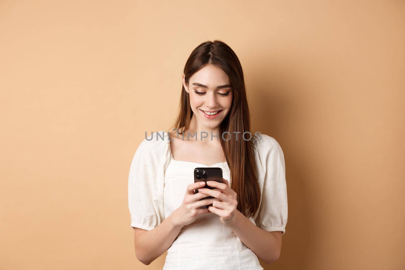 Young woman texting message on mobile phone, smiling and reading smartphone screen, standing on beige background.