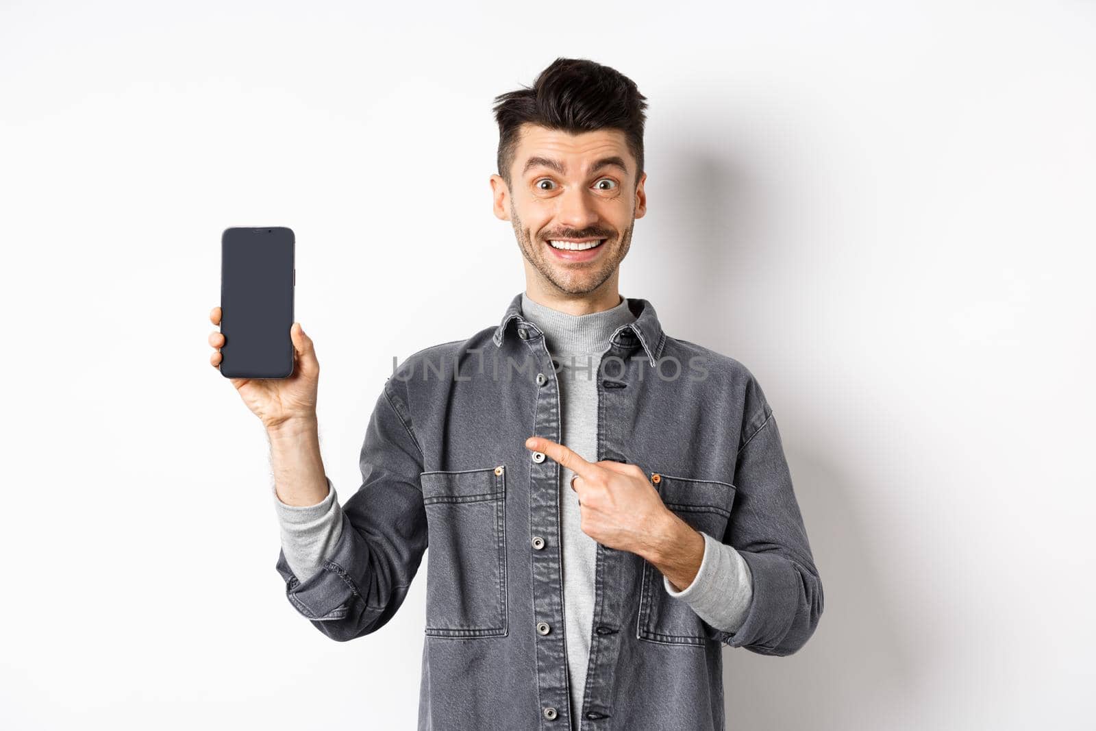 Handsome smiling man pointing finger at smartphone screen, showing mobile phone display with happy face, standing against white background by Benzoix