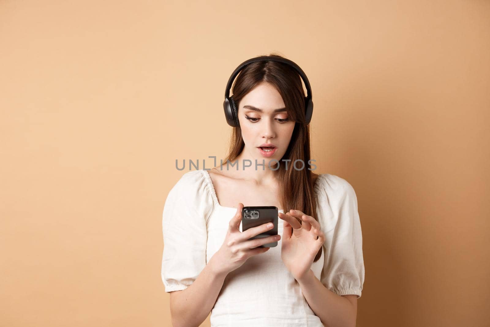 Excited girl listen music in headphones and looking at smartphone amazed, reading awesome news, found cool playlist, standing on beige background by Benzoix