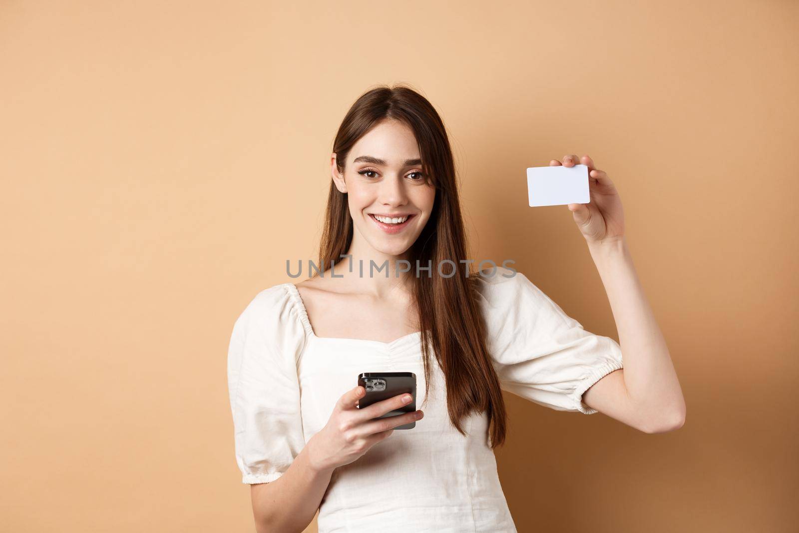 E-commerce concept. Smiling woman showing plastic credit card while shopping online on mobile phone, standing against beige background.