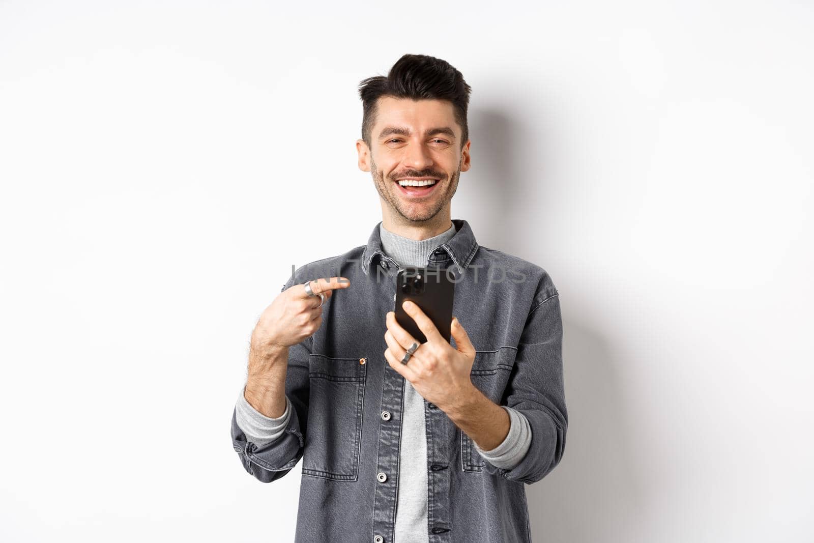 Happy handsome caucasian man with moustache pointing finger at smartphone, smiling and showing good deal online on mobile phone, standing pleased against white background by Benzoix