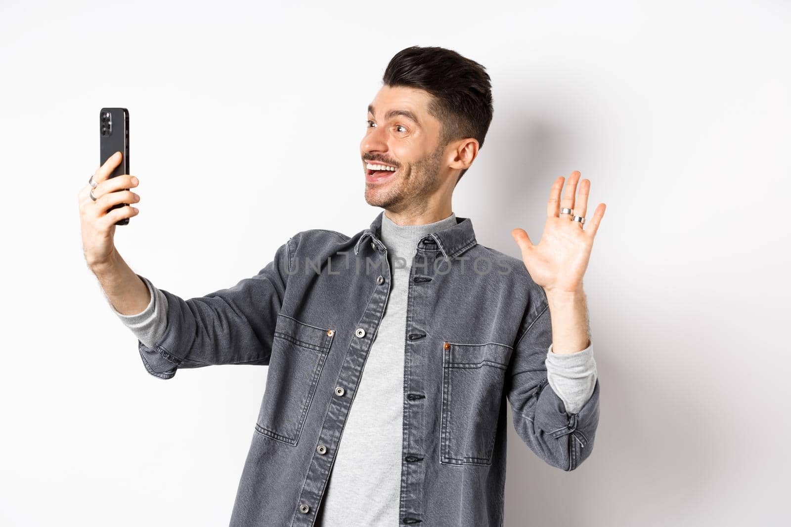 Young man video chatting on mobile phone, waiving hand at smartphone camera, greeting friend online, standing on white background.