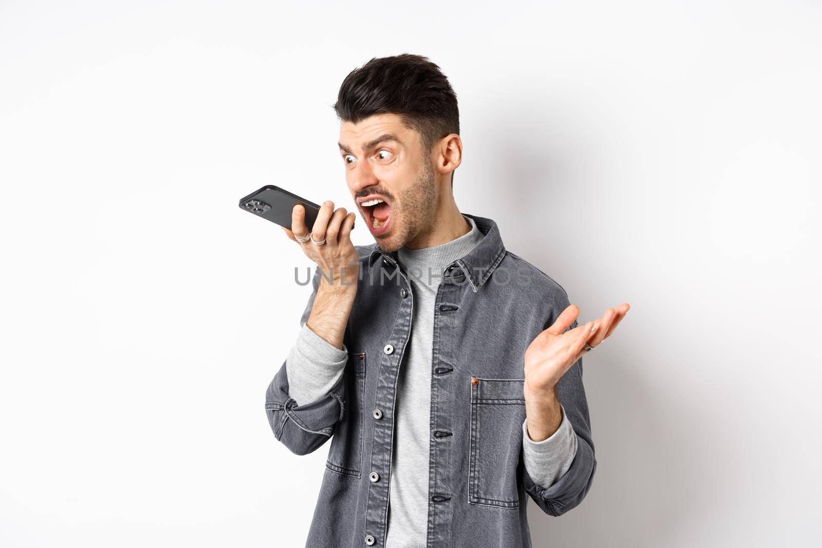 Angry guy shouting at speakerphone, scream on mobile phone, leaving angry voice message, standing against white background.