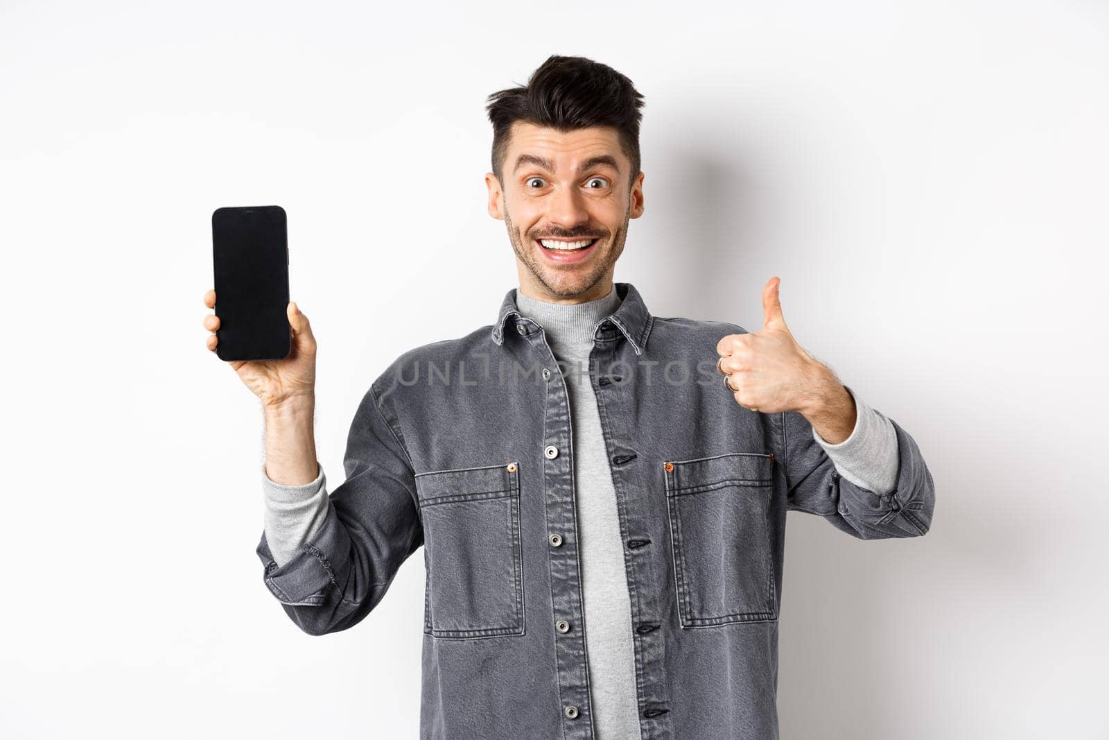 Cheerful stylish man showing empty smartphone screen and thumbs-up, concept of online shopping and e-commerce by Benzoix