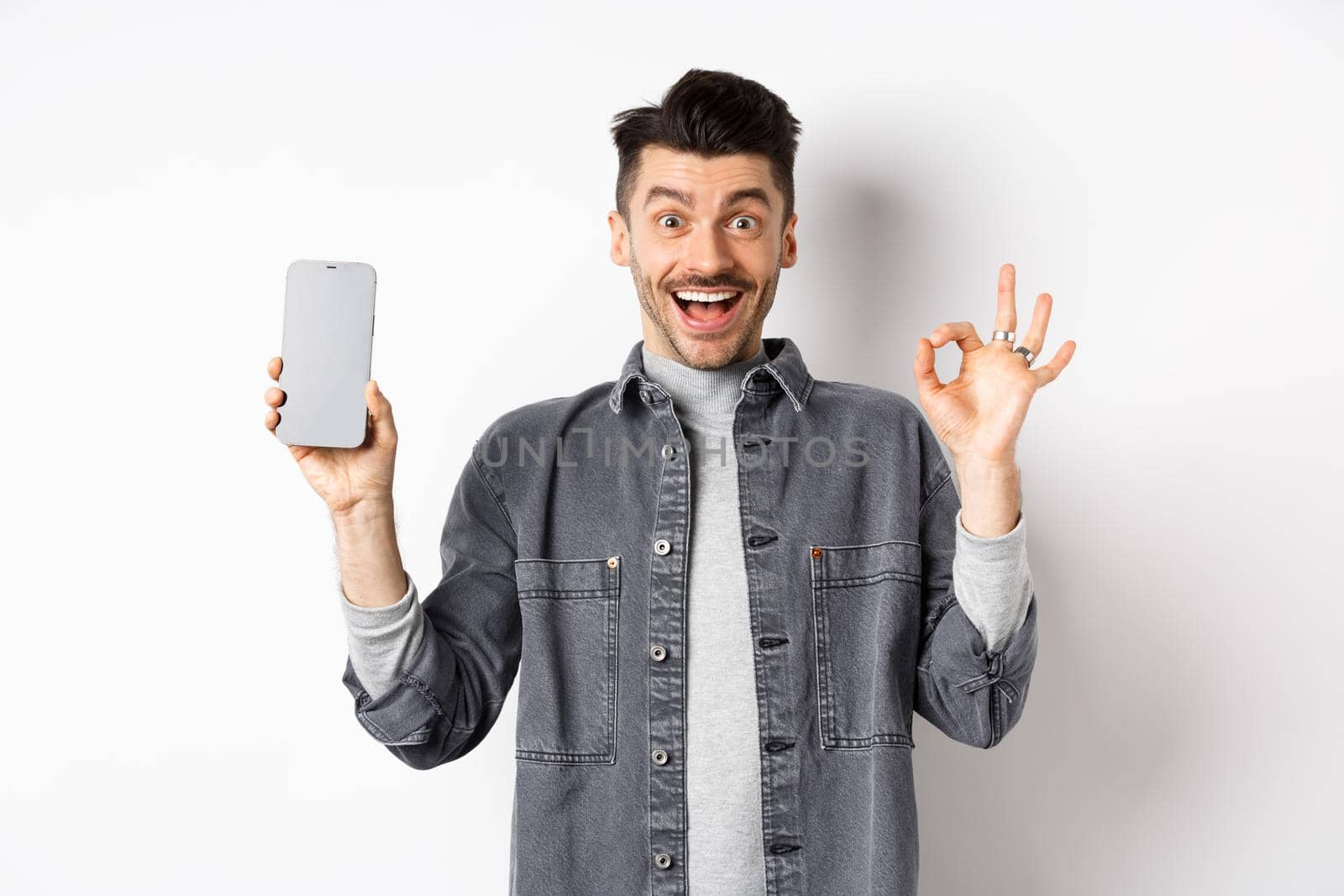 Excited smiling guy showing empty smartphone screen with okay gesture, recommending app or shopping offer, standing on white background.