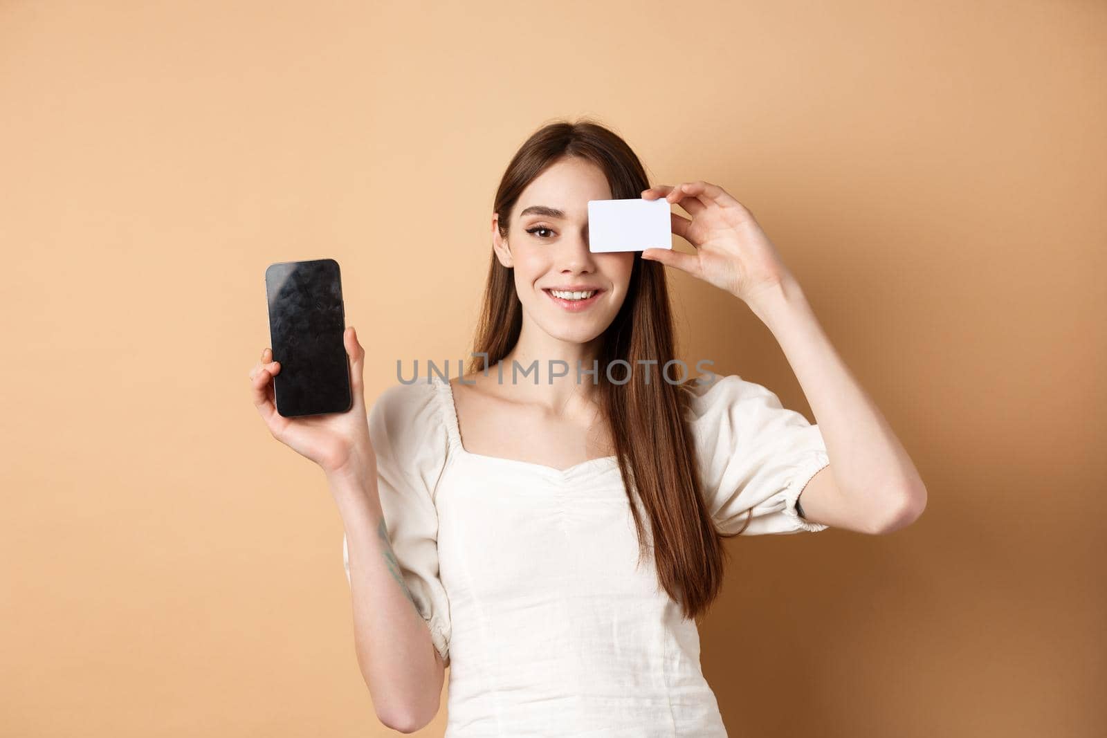 Cheerful young woman showing plastic credit card and empty mobile phone screen, smiling pleased at camera, standing on beige background.