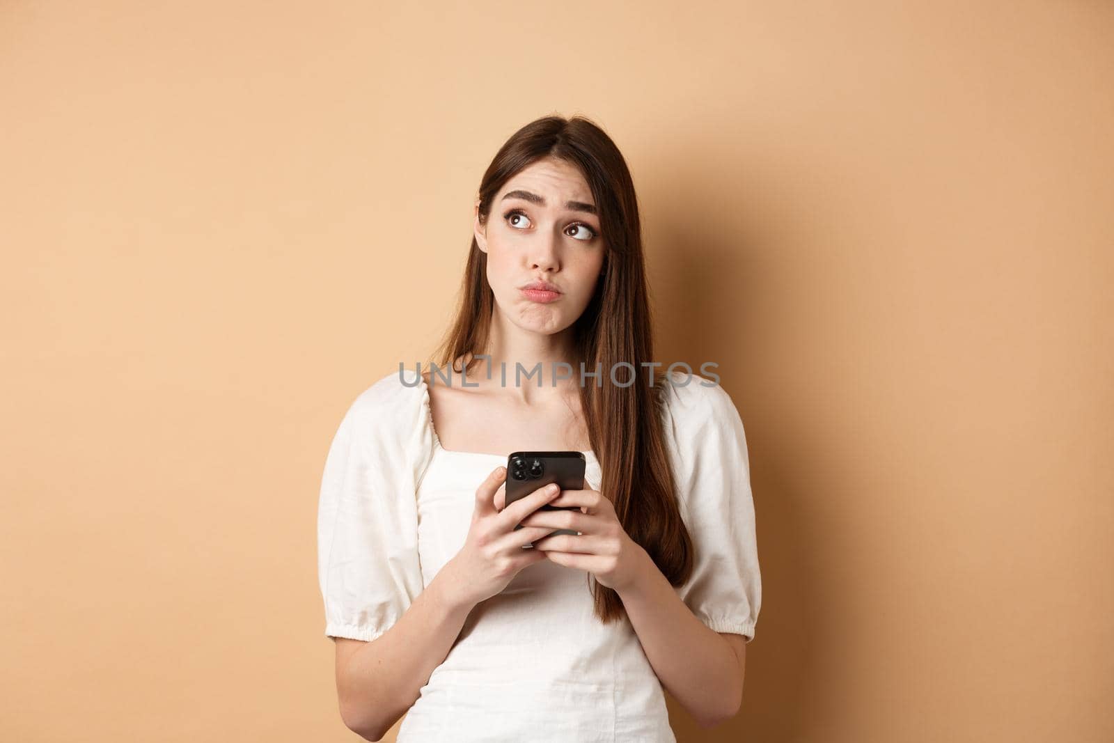 Confused young woman thinking after reading news on mobile phone, looking at upper left corner hesitant, standing on beige background by Benzoix