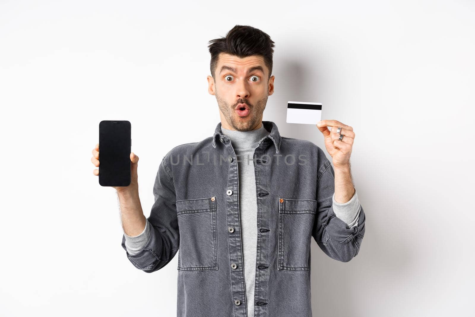 Excited handsome man showing plastic credit card and mobile phone screen, standing on white background.