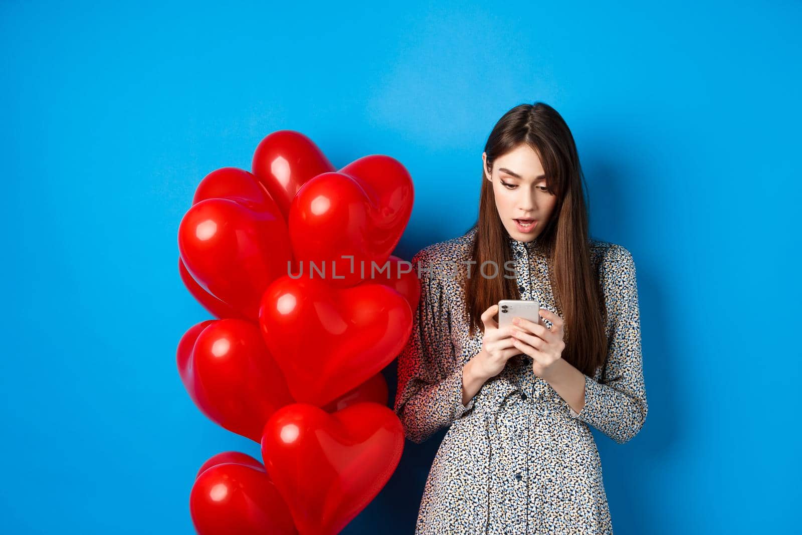 Valentines day. Portrait of young woman standing near red romantic balloons, looking surprised at smartphone screen, blue background by Benzoix