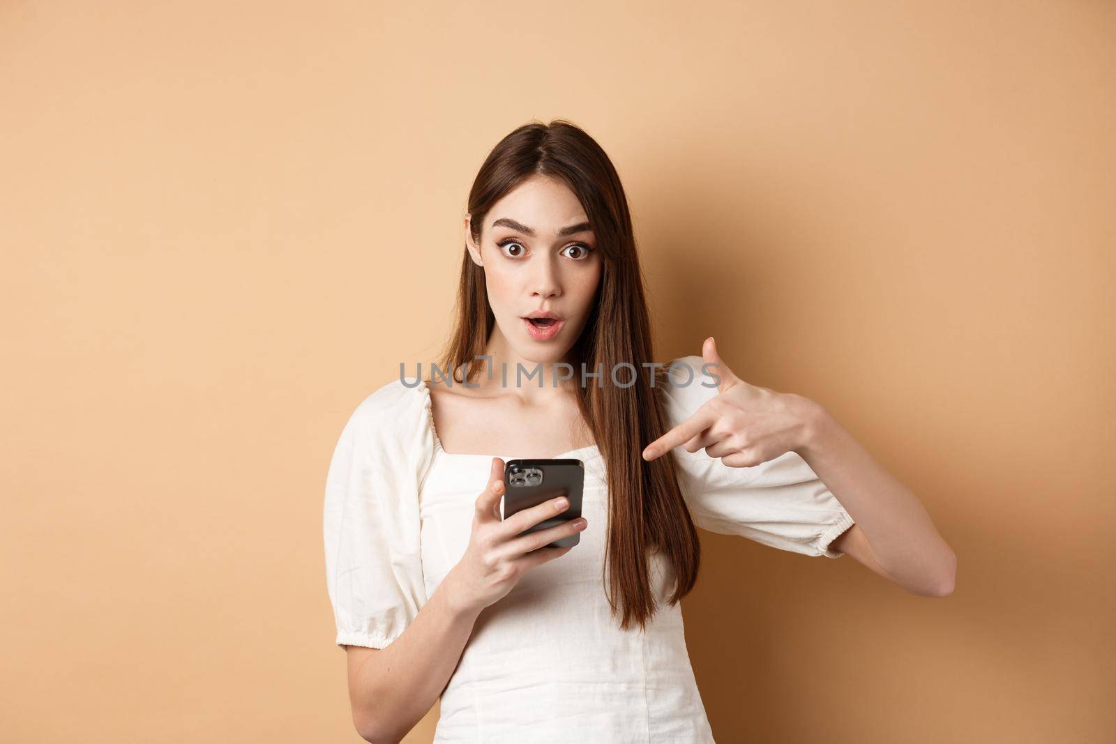 Online shopping concept. Excited girl pointing at smartphone screen and looking at camera amazed, standing against beige background.