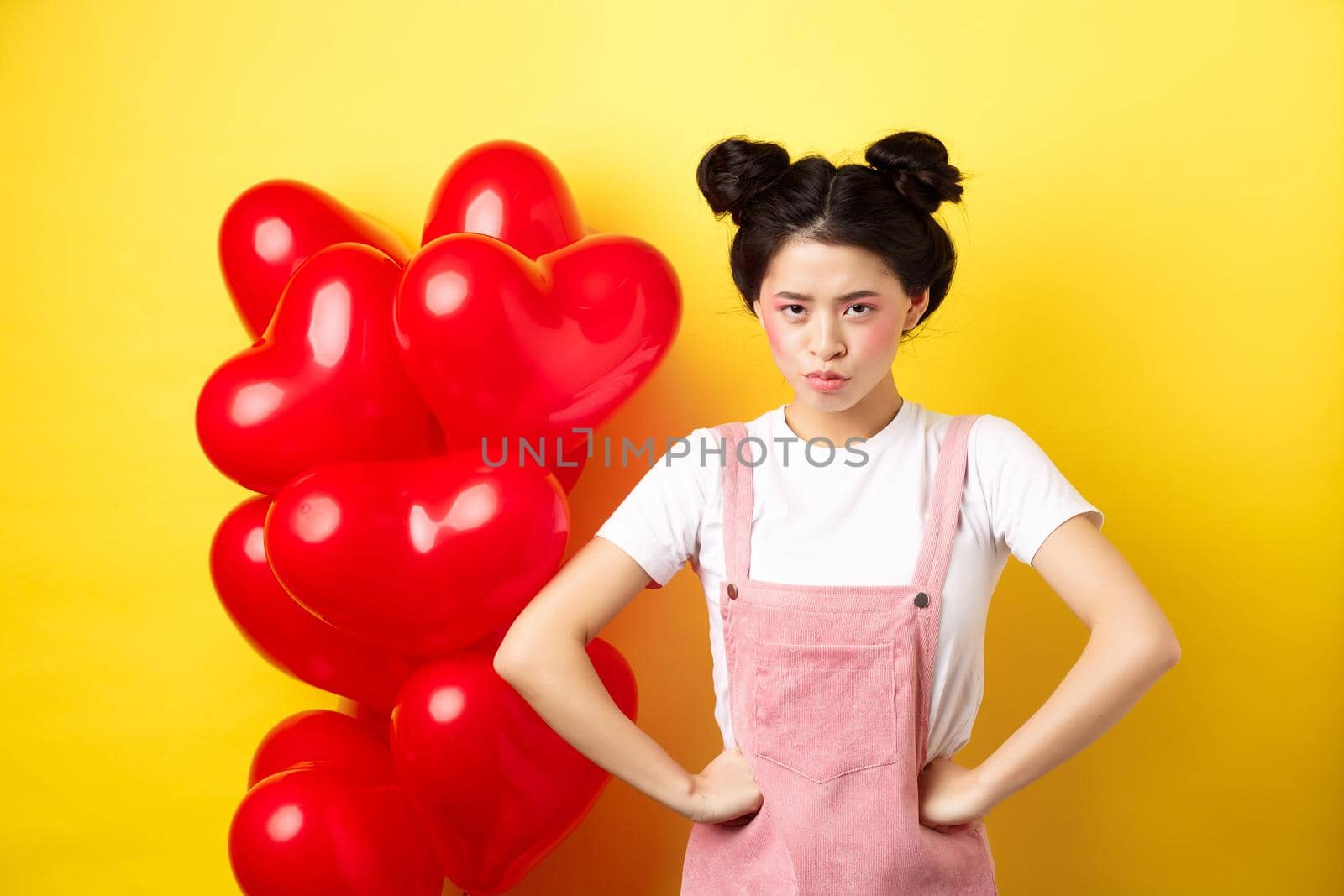 Valentines day and relationship concept. Disappointed asian girlfriend sulking, looking offended at camera, mad at boyfriend, standing near heart balloons, yellow background by Benzoix