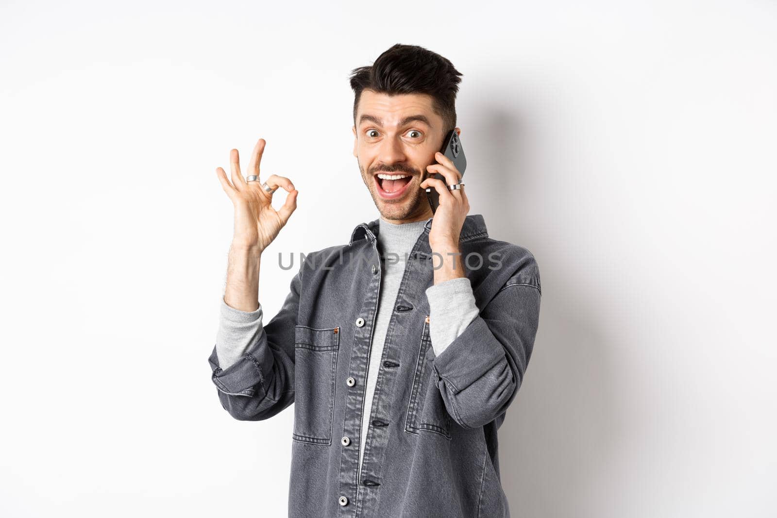 Excited young man talking on phone and showing okay gesture, smiling satisfied, standing against white background.