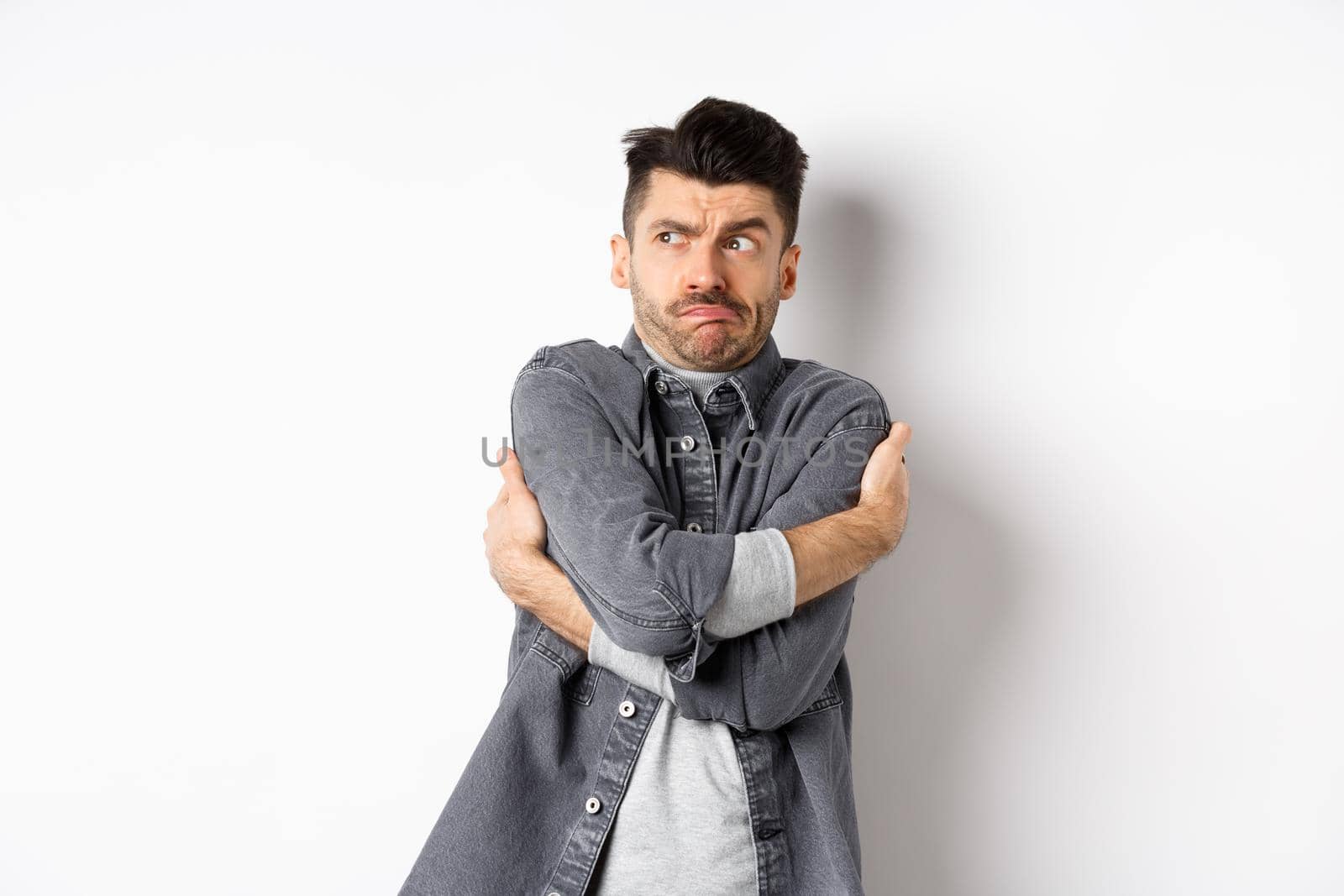 Displeased young man feeling discomfort, hugging himself and staring left at something embarrassing or exposing, standing uncomfortable on white background.