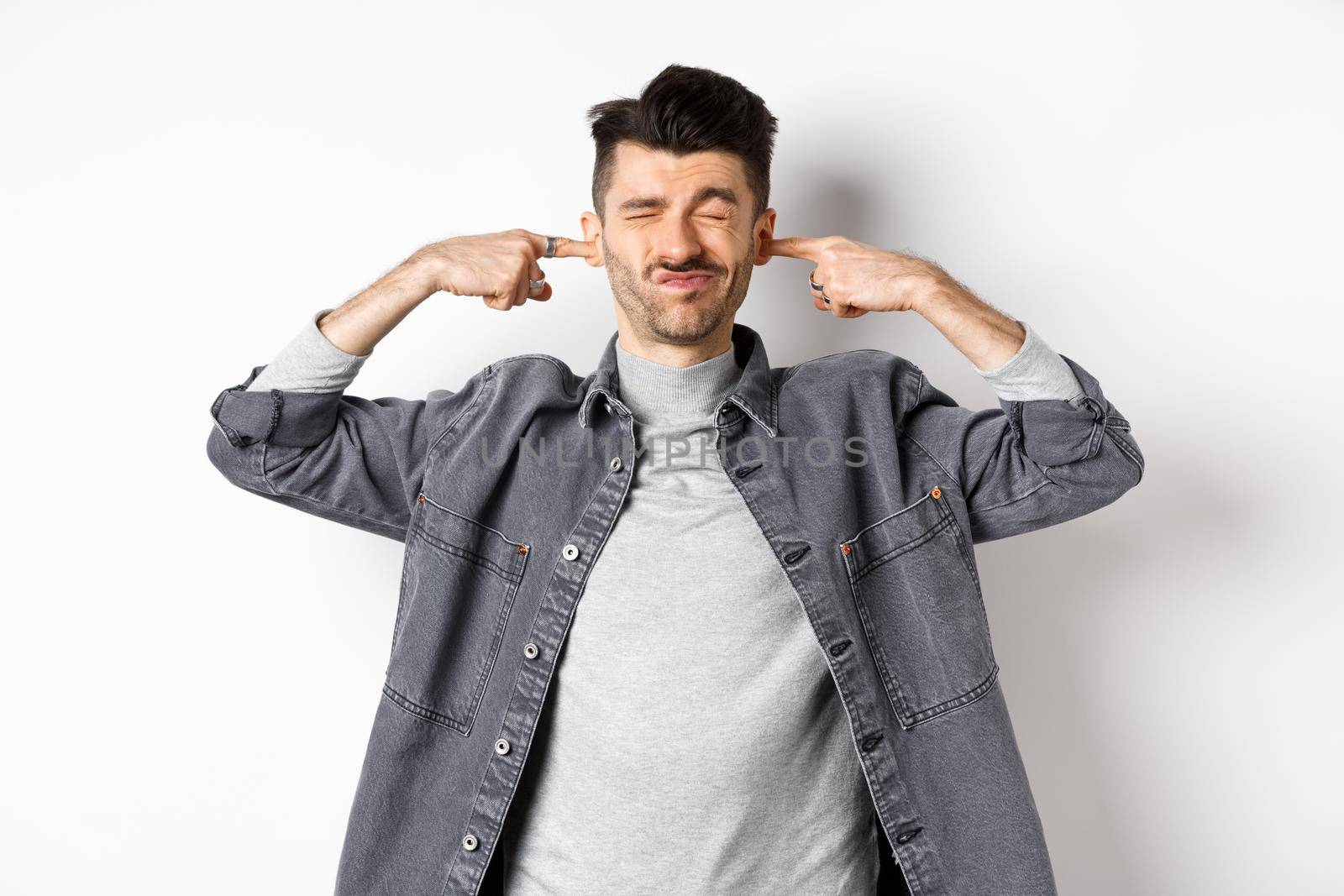 Annoyed young man plug ears with fingers and grimacing from loud noise, disturbed by noisy neighbours, standing at loud place on white background.