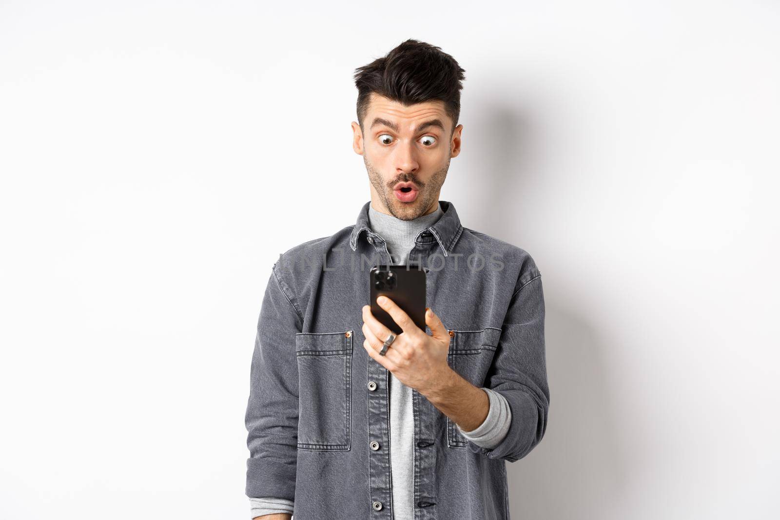 Man look surprised at smartphone screen, receive exciting news on phone, standing against white background.