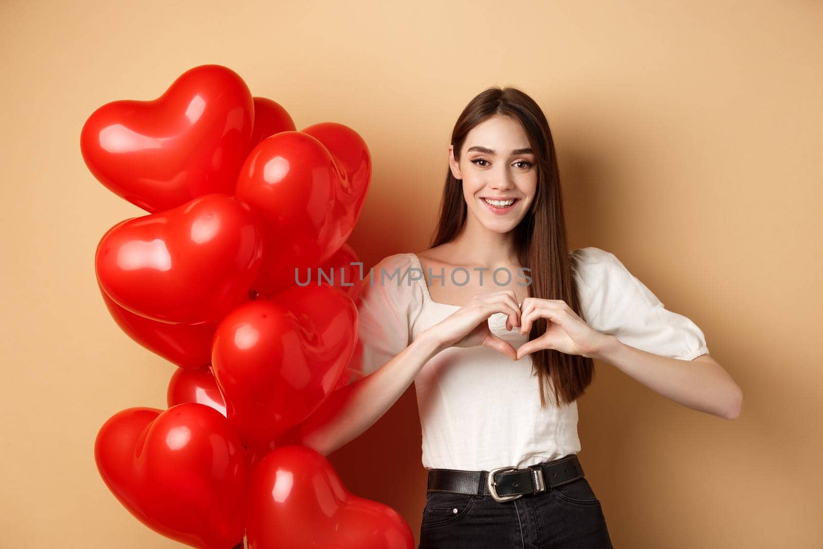 Lovely caucasian girl express love and tenderness, showing heart gesture and smiling, standing near Valentines day balloons, beige background by Benzoix